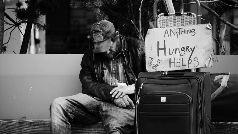 A homeless man sitting next to a sign and his luggage